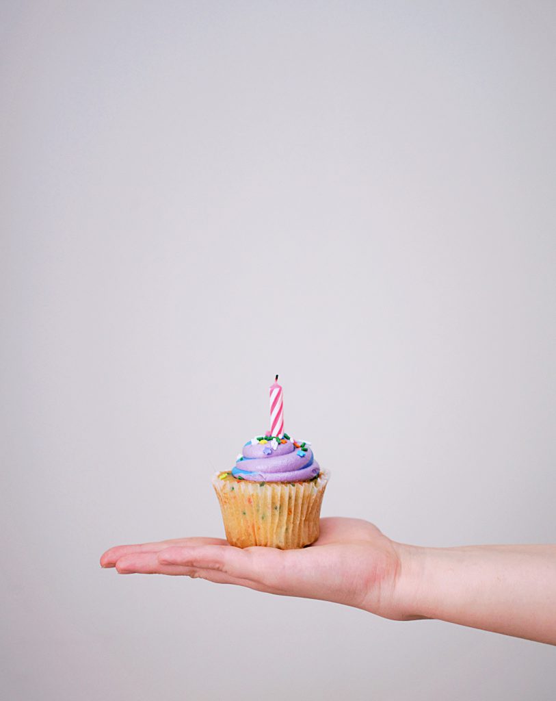 A mão de uma pessoa branca segurando um cupcake confeitado com uma vela.