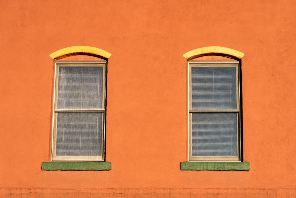 Foto de uma parede laranja com duas janelas retangulares, peitoril verde e persianas do lado de dentro.